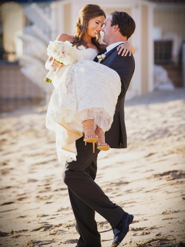 A groom in a black suit carries a bride in a white wedding dress and heels on a sandy Orlando beach. The bride holds a bouquet of flowers, and they are both smiling at each other.